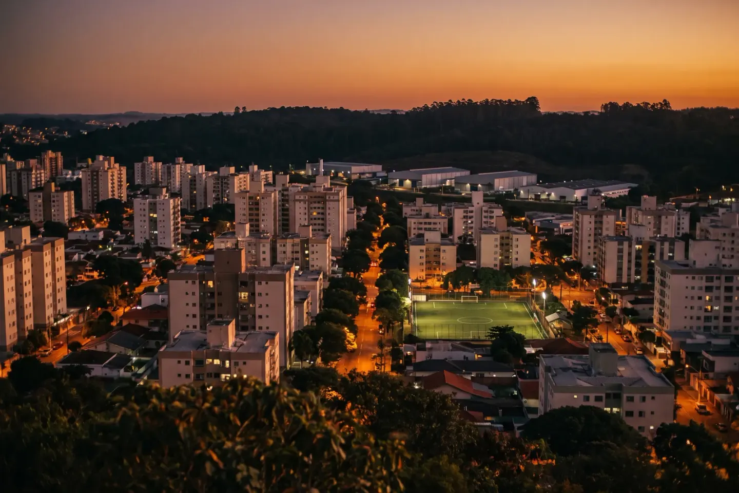 Vista do alto de uma cidade adensada ao entardecer: prédios de poucos andares, avenidas arborizadas e um campo de futebol iluminado.