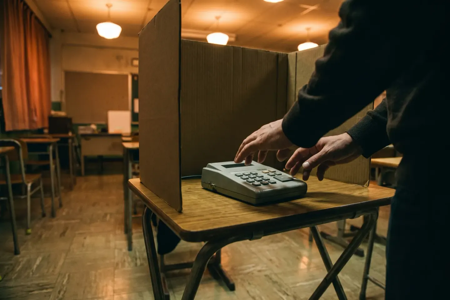 Mãos de um eleitor digitam no teclado de uma urna apoiada numa carteira escolar, atrás de uma cabina de papelão.